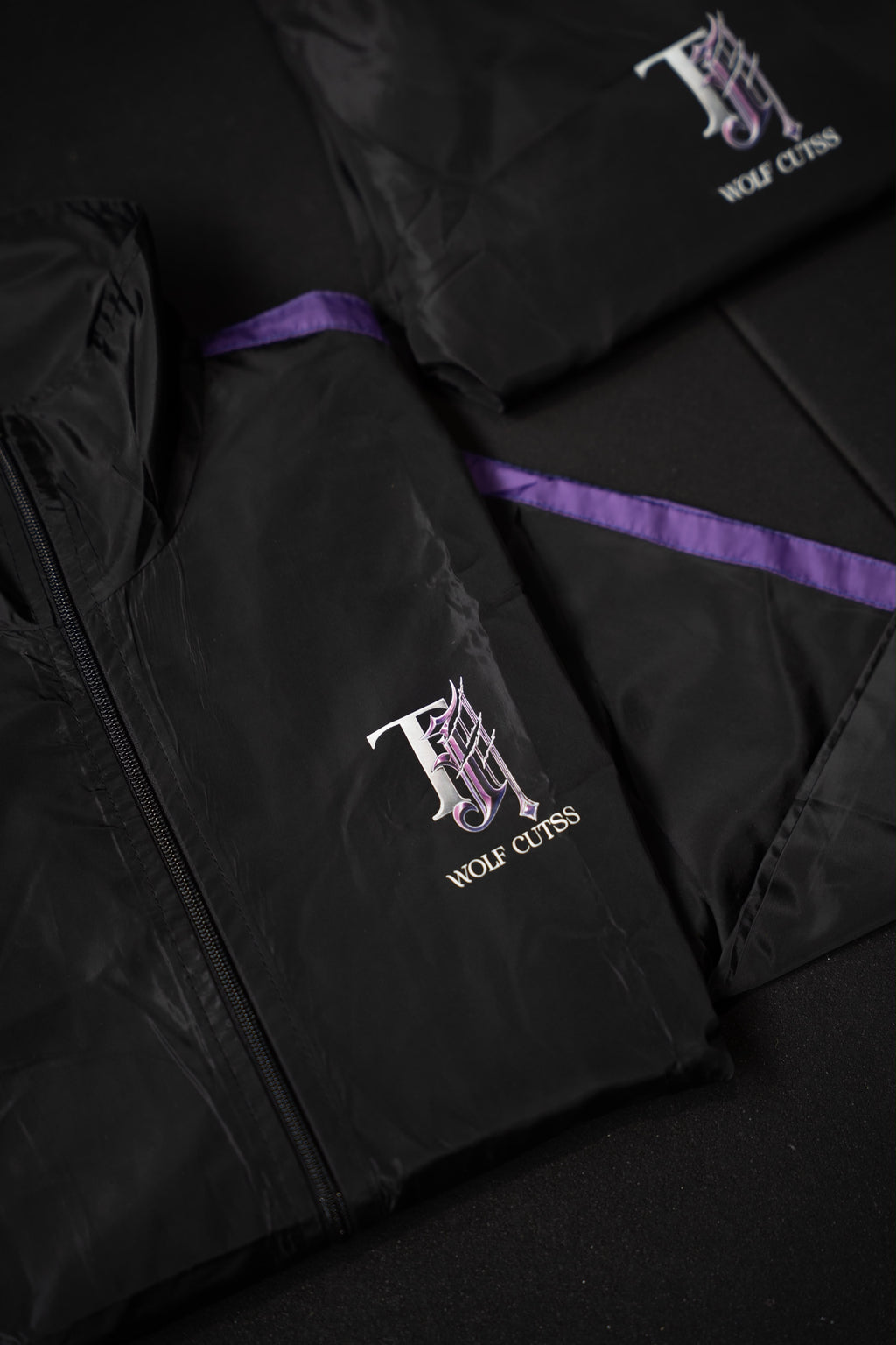 Black TJ Wolf Cutss tracksuit jacket and pants folded flat, showing purple side stripe and chest logo