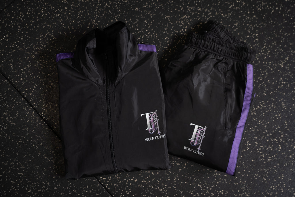 Black TJ Wolf Cutss tracksuit jacket and pants laid out on dark gym flooring