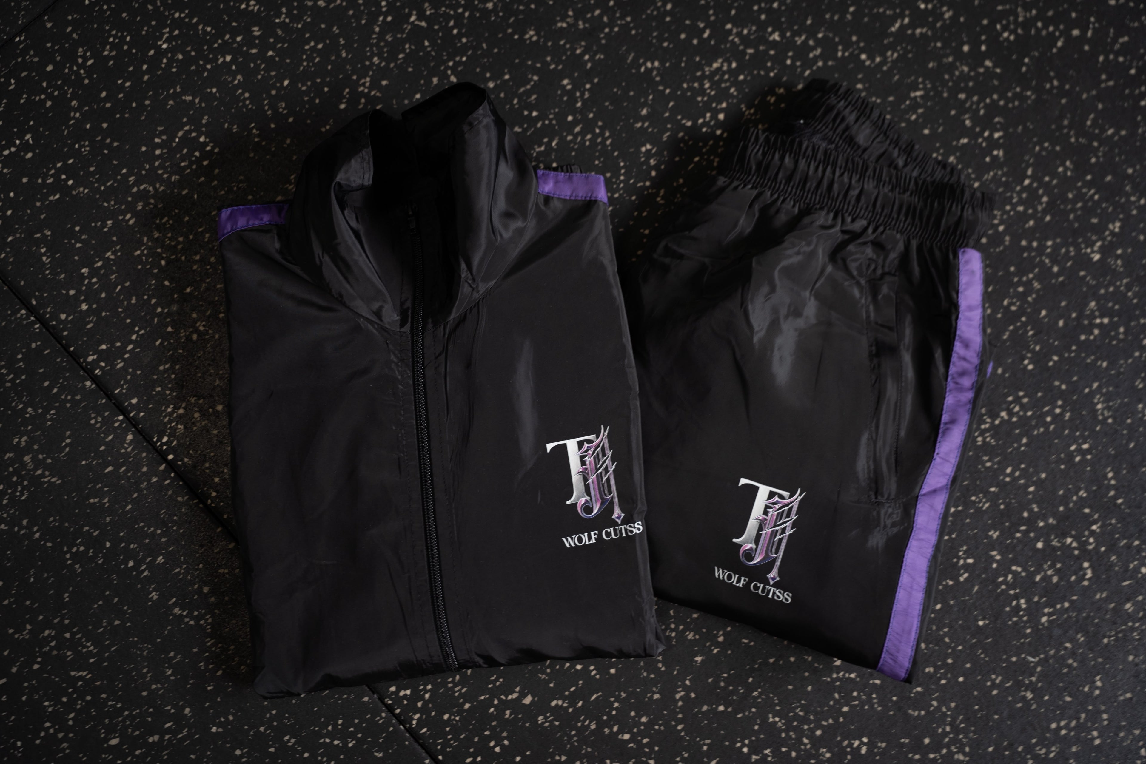 Black TJ Wolf Cutss tracksuit jacket and pants laid out on dark gym flooring