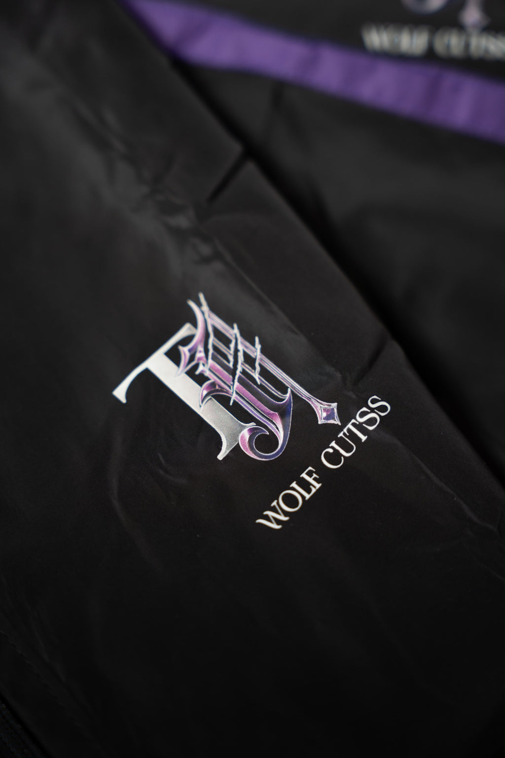 Close-up of the purple and silver TJ Wolf Cutss gothic logo on black tracksuit fabric