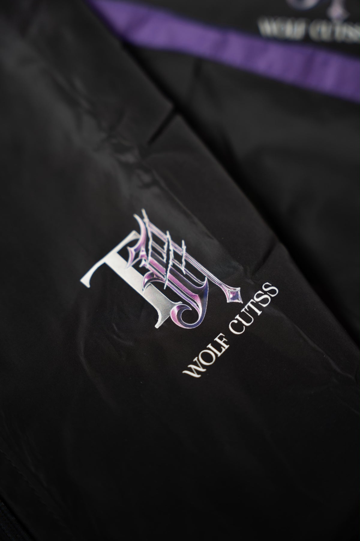 Close-up of the purple and silver TJ Wolf Cutss gothic logo on black tracksuit fabric