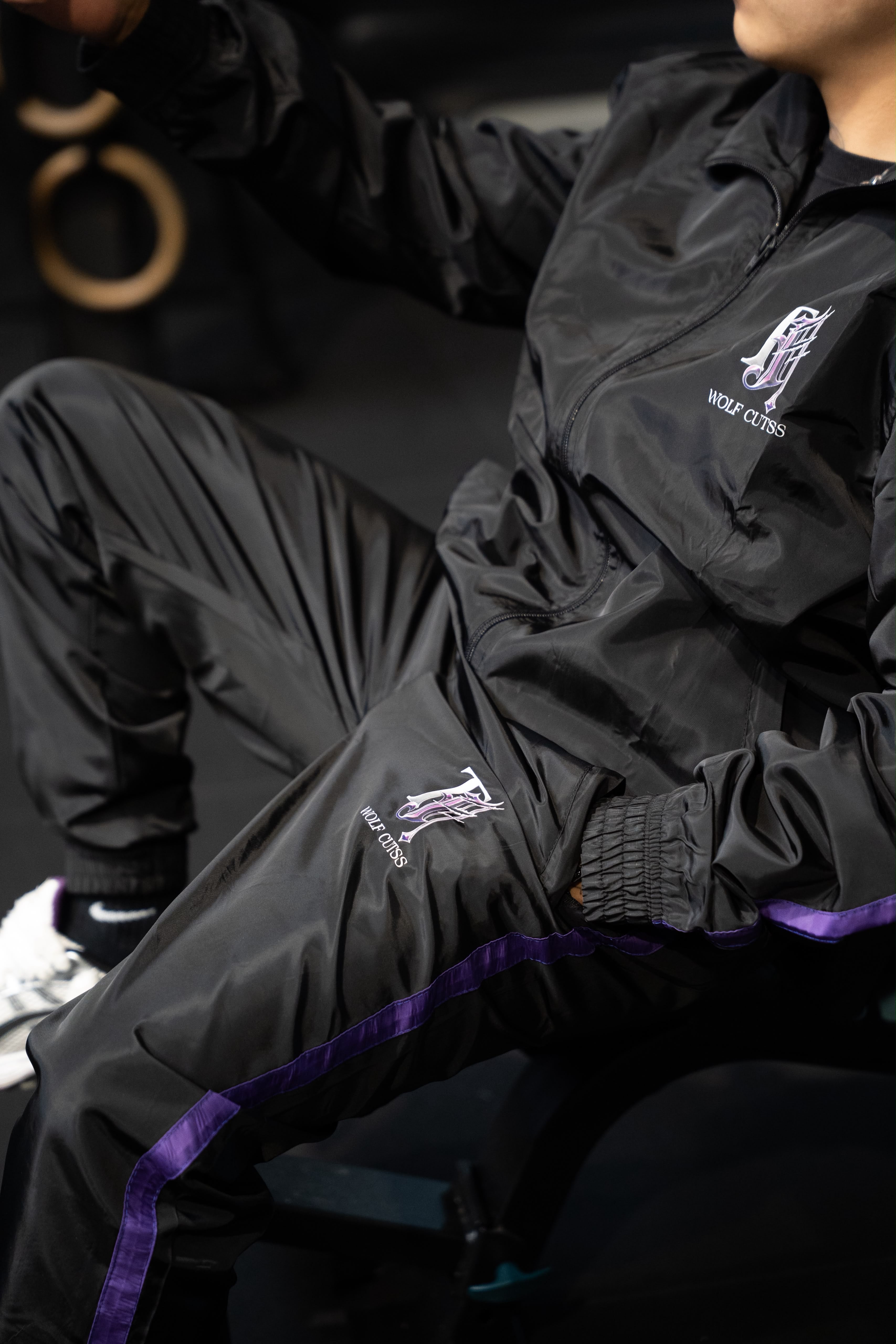Model seated wearing the black TJ Wolf Cutss tracksuit with purple stripe down the leg