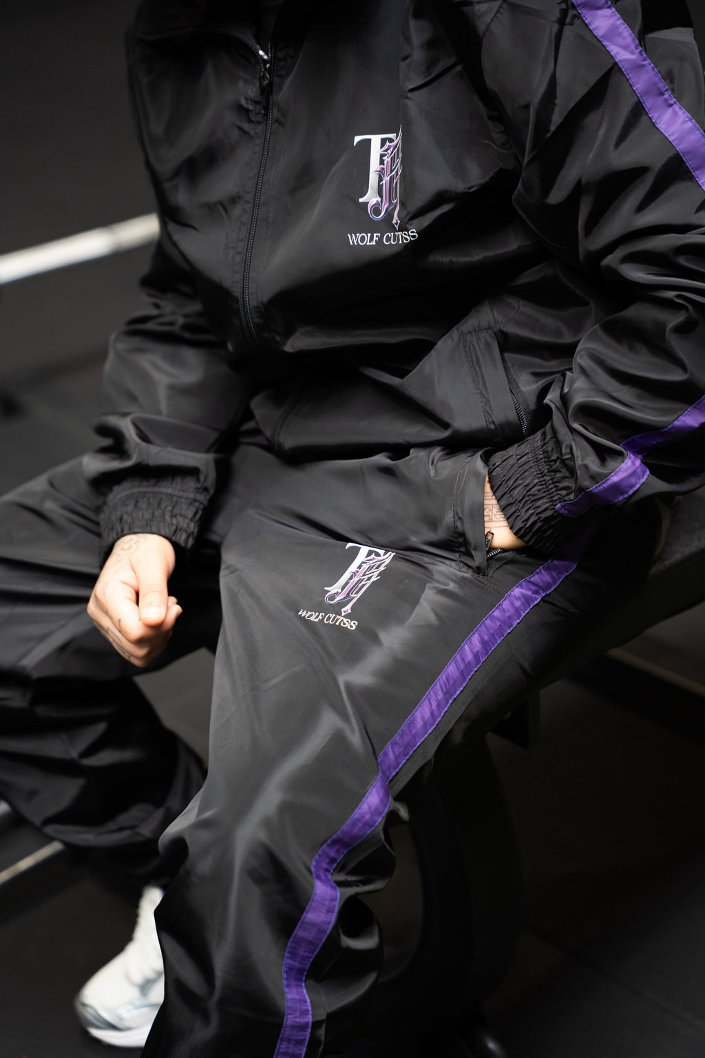 Model sitting in the black TJ Wolf Cutss tracksuit, purple side stripe visible on the trousers