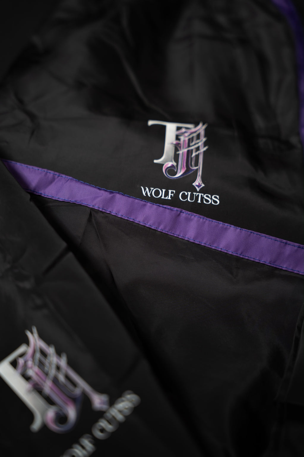 Detail of the TJ Wolf Cutss chest logo and purple stripe on the black tracksuit jacket
