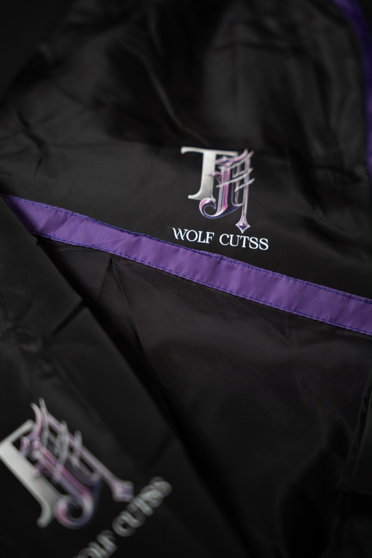Detail of the TJ Wolf Cutss chest logo and purple stripe on the black tracksuit jacket